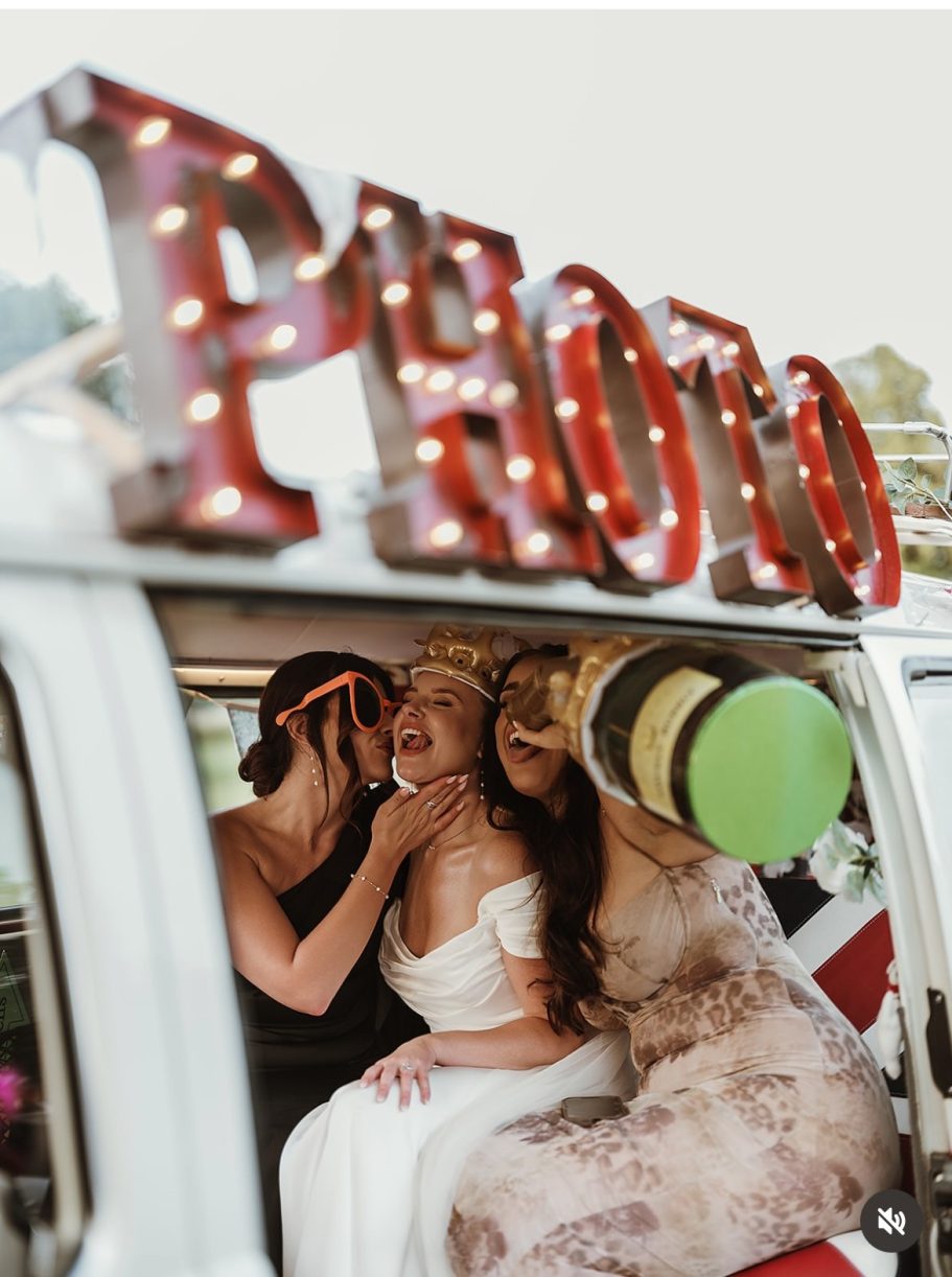 VW campervan photobooth Three women in wedding attire pose playfully inside a vintage van with a 'PHOTO' sign.