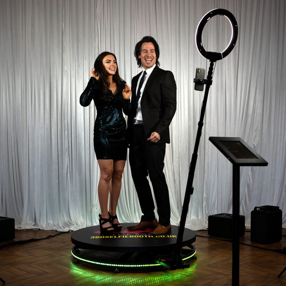 360 photo booth hire in Kent A woman in a black dress and a man in a suit pose on a circular platform with a ring light.